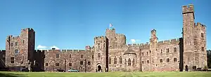 A sandstone castle-like building with a tower slightly to the left of centre. The walls are battlemented and in the foreground is a grassed area