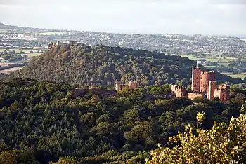 A wooded area with a sandstone castle on the right. Beyond is another wooded hill with the ruins of a castle on its top. In the far distance are fields and houses.