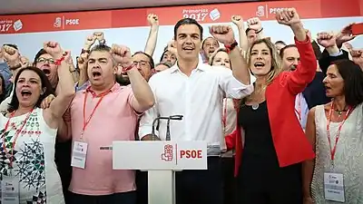 Pedro Sánchez wearing a white shirt and cheering with multiple people