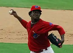 A right-handed man in a red baseball jersey and red baseball cap throws a baseball. His jersey and cap have a teal-colored shark on the front, and he is wearing a black baseball glove on his left hand.