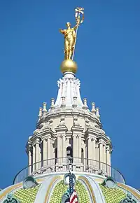 A stone, columned cupola atop a green, tiled-dome. The cupola is topped with a gold globe, upon which stands a gold statue with an outstretched arm and holding a staff.