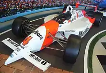 A red and white racing car with sponsors logos adorning it and the number 4 on the front wing sitting in a museum