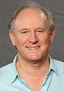 Portrait photograph of Peter Davison wearing an open‑necked, cyan shirt.