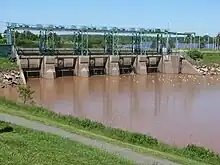 A concrete and steel structure crosses the river. The water is brown on the near side of the structure, and blue on the far side