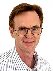 A man with glasses and white-and-black-striped color shirt stands in front of a white background.