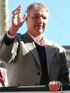 A gray-haired man stands in the back of a truck. He is wearing a patterned jacket, a pink shirt and a dark sweater vest. His right hand is raised.