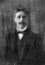 Head and torso of standing middle-aged white man, in a dark suit, with full head of wavy dark hair, looking to the side. He has a neat, medium-sized moustache