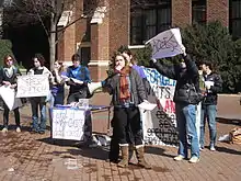 A young woman speaks into the microphone of a bullhorn in front of a folding table while others around her hold signs with the words "ACCESS" and "FREE SPEACH" crossed out.