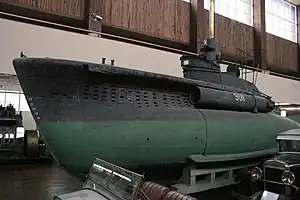 a colour photograph of a small submarine with dark green paint on the lower hull and dark gray paint on the upper hull. The submarine is located in a large indoor space and located among other objects.