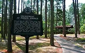 Historic marker reading "Engagement at Poison Springs" in foreground of a forested area with rustic wooden pavilion in the background.