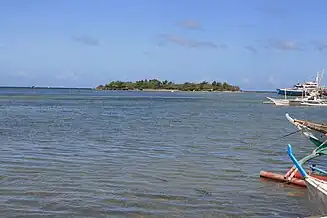 Small low green island in blue sea, at about 600 m, with several palm trees on top.  To right prows of four bangkas – local boats for fishing or diving.