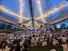 Pomona alumni eating on Marston Quad at round blue tables with plastic chairs under a translucent tarp with string lights