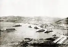 A black and white shot of several ships lying submerged underwater in various states of capsizing, a white building is shown in the right corner of the foreground.