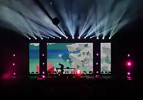 On a distant stage, many lights shine outward, and Porter Robinson is silhouetted against a screen depicting the sky and floating cuboidal structures.