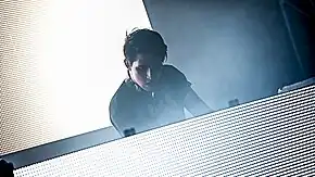 Porter Robinson stands at a table with a brightly-lit screen; another screen is behind him