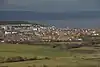 Multiple buildings including terraces, detached houses and blocks of flats. In the foreground are fields and in the background water and then hills.