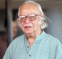 Close up of an old semi bald man wearing broad black framed glasses.