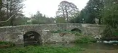 Gray stone bridge with two arches over water. The central pillar is on a small island. Trees to the left and right and behind the bridge.