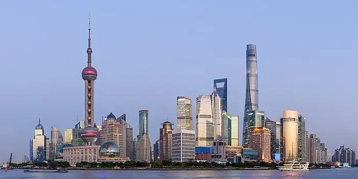 A group of skyscrapers and towers, seen from across a river. At the left is one consisting of a sphere on concrete supports topped by a long spike; in the center are smaller buildings, one a bright gold color, gradually rising to the tallest one at right, still under construction