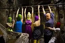  A group of 6 women wearing ski masks and multicolored clothes in a small, grubby brick space. Four of them hang by their arms from a bar, one flexes her muscles, and another grabs one of the hanging women around the waist.