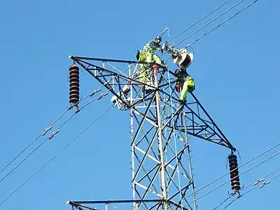 Cable riggers atop a pylon adding a fibre optic data cable wound around the top tower stay cable. The cable (SkyWrap) is wound on by a traveling machine, which rotates a cable drum around the support cable as it goes. This travels under its own power from tower to tower, where it is dismantled and hoisted across to the opposite side. In the picture, the motor unit has been moved across but the cable drum is still on the arrival side.