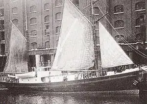 A ship with a black hull and white upper parts, three sails raised, in dock alongside a high multi-windowed warehouse building