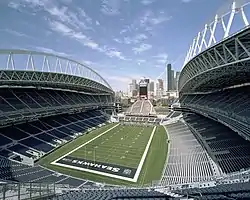 The interior of a stadium from the upper tier behind the south end zone during the day. The end zones and seating sections are colored blue. At the north end is a smaller seating area at the base of a tower. Several high-rise office buildings are in the distance.