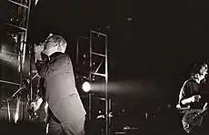 A black-and-white photograph of Michael Stipe and Peter Buck performing on stage with spotlights on them. Stipe is to the left singing into a microphone, wearing a three-piece suit, he has bleach-blond hair and is obscuring Mike Mills, whose bass guitar is visible from behind him. Peter Buck is playing guitar and wearing a button-up pattern shirt behind Stipe to the photograph's right with a sneer on his face.