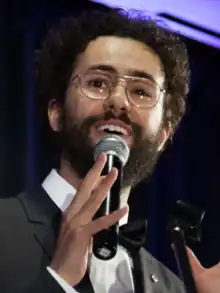 Youssef holding a microphone and smiling while wearing a tuxedo