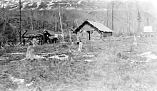 A black and white photo of two log cabins in a lightly wooded area.