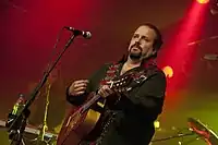 Singer Raul Malo, playing an acoustic guitar against a red background.