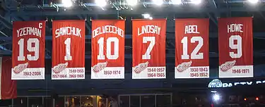 A row of six red banners.  The banners, from left to right, read "Yzerman 19" "Sawchuk 1" "Delvecchio 10" "Lindsay 7" "Abel 12" "Howe 9". The Yzerman banner has a small "C" at the top right corner.