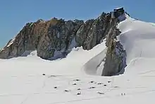 mountain hut on rock outcrop with climbers camping on nearby glacier