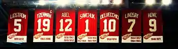 The banners of seven retired numbers.  The banners, from left to right, read "Lidstrom 5" "Yzerman 19" "Sawchuk 1" "Delvecchio 10" "Lindsay 7" "Abel 12" "Howe 9". The Yzerman banner has a small "C" at the top right corner.