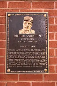 A dark bronze plaque with a man's face at the top and gold writing beneath, mounted on a brick wall