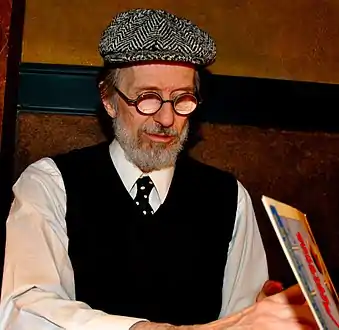 An elderly man with a white beard, round glasses, a beret-like hat, a dark vest, and a necktie. He faces down right, looking into an open book.