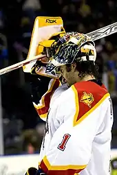 An ice hockey goaltender removing his mask. He wears a white and orange jersey with the number "1" on his elbow.