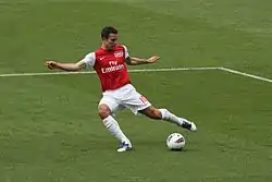 A coloured photograph of a footballer wearing a red-and-white football shirt with the team logo. He is positioning himself to kick the ball with his left foot.