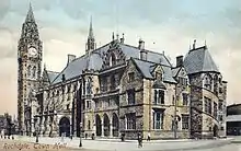 A large, ornate and angular taupe-coloured building on a sunny day. The building fills the image with the exception of flat, grey pavement at the building's frontage, which is dotted with people. The multi-storey building has many small slitted triangular and rectangular bays and windows across its façade, except for a dark arched entrance at ground-level. At the far end of the building is a huge clock tower topped with a triangular spire standing tall at around 15 to 20 times the height of an average adult human.