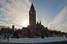 A fine and ornate building with a huge clock tower fills the central part of the image. It appears directly in front of the sun giving it a dark silhouette-like setting. The scene is marked by white snow.