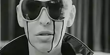 A woman's front facial profile expressing an emotion of shock as blood leaks from her eye behind her vintage sunglasses