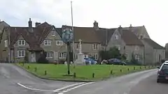 Street scene. Triangular area of grass with village sign on wooden post and stone cross behind. Stone houses with tiled roofs in the background.