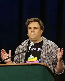 A Caucasian man with average length brown hair wearing an open casual shirt and a T-shirt gestures while making a speech from a podium.