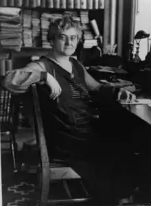 Black and white photograph of a woman in a library, seated at a desk with her arm draped over a chair, holding a pencil in her right hand