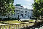 A white stone library with a green dome on top and two lions in front surrounded by grass and bushes