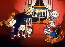 A cartoon of an elderly man lighting a Menorah. He is bald and wearing a Kippah. At his feet are three toddlers; two are on their hands and knees, the other is standing. To their right are two infants sitting on a large brown dog. One infant is bald and wearing a nappy; the other is wearing a t-shirt and shorts.