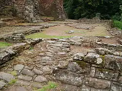 Ruined foundations of buildings, some patches of ground covered in cobblestones; at the far and near ends of the foundations the stonework is built up to around a metre tall; a castle wall can be seen in the background left.