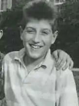 A black and white photo of a Caucasian teenaged male. He is wearing a light colored polo with stripe's, button's by the neckline and a neck collar. Another person's ear can be seen with his arm on the shoulder of the teenage male.