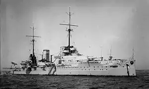 Right front oblique view of a white-painted warship at anchor. The ship has two masts, two funnels, and two gun turrets visible.