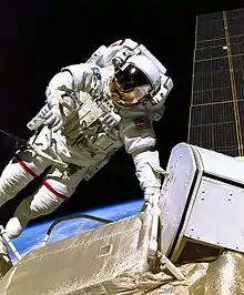 An astronaut in a white spacesuit with a red, white, and blue flag on the left shoulder in the foreground with a black background